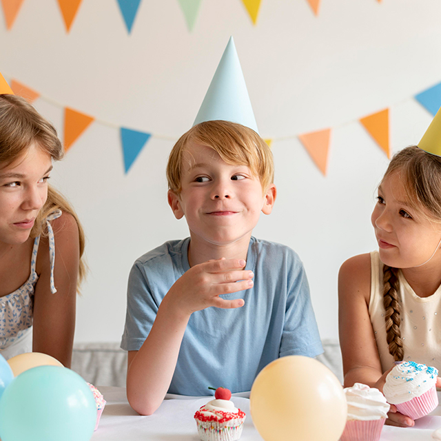 Drei glückliche Kinder mit Geburtstagshüten und Luftballons sitzen am Tisch und essen Muffins